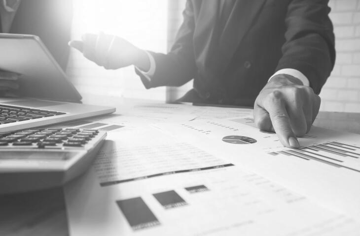 Two business people sit at a desk analysing financial charts and graphs. One man points to a specific data point on the document in front of them, while a laptop and calculator sit nearby to aid in their analysis and discussion.